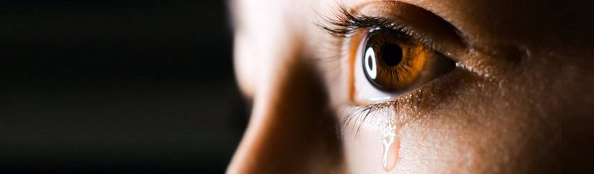 A close up of someone with hazel eyes crying on a black background.