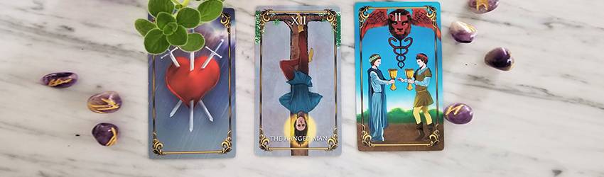 Three Tarot cards from the Astrology Answers Master Tarot Deck: The Hanged Man, 3 of Swords, and 2 of Cups. These represent the themes of July 2020 in this Tarotscope reading.
