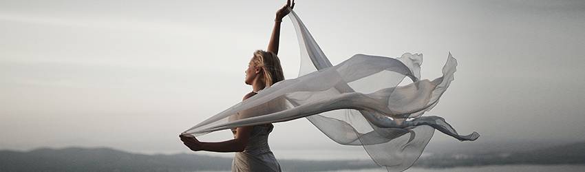 A blonde woman wearing a white gown holds up white gauzy fabric behind her, which appears to be blowing in the wind.