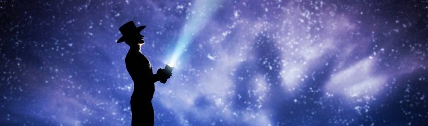 A shadowed figure stands, holding a flashlight up to the night sky. The expansive sky is illuminated by the white stars in shades of blue, purple, and black.