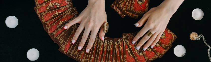Hands spread over the backs of red and gold Tarot cards. The table they are on also has candles and a pendant.