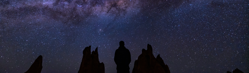 A silhouetted figure stands amongst the stars of the night sky, basked in colors of dark purple and blue.