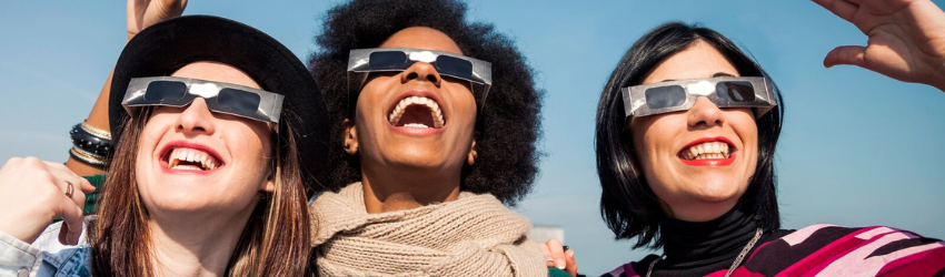 Three women wear special eclipse sunglasses and look into the sky excitedly.